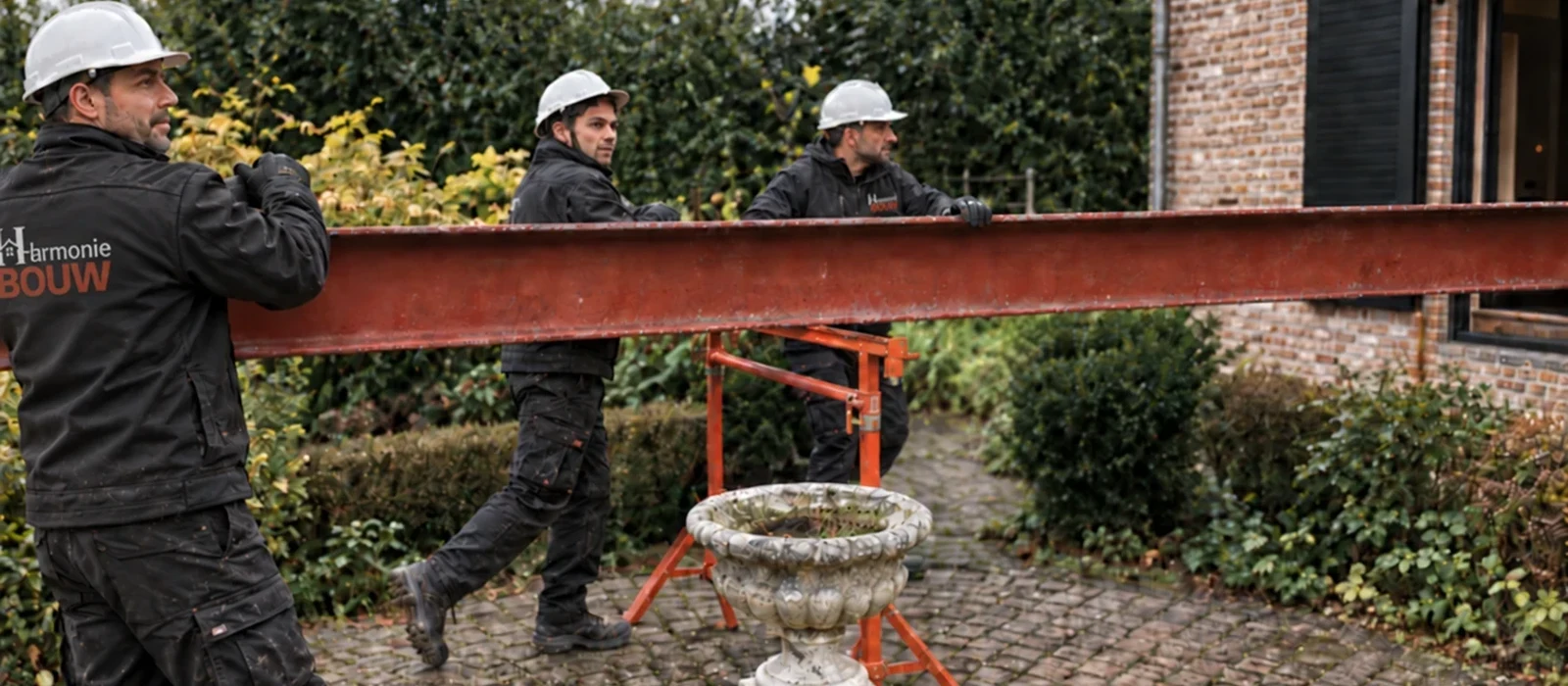 Vier vaklui van Harmonie Bouw met helm en bedrijfskledij dragen samen een stalen ligger naar de werf