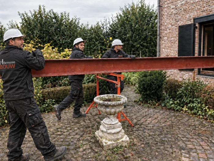 Drie vaklui van Harmonie Bouw met helm en bedrijfskledij dragen een stalen ligger naar de werf
